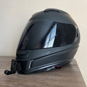 May include: A black motorcycle helmet with a clear visor and a black camera mount attached to the bottom. The helmet is sitting on a wooden surface.