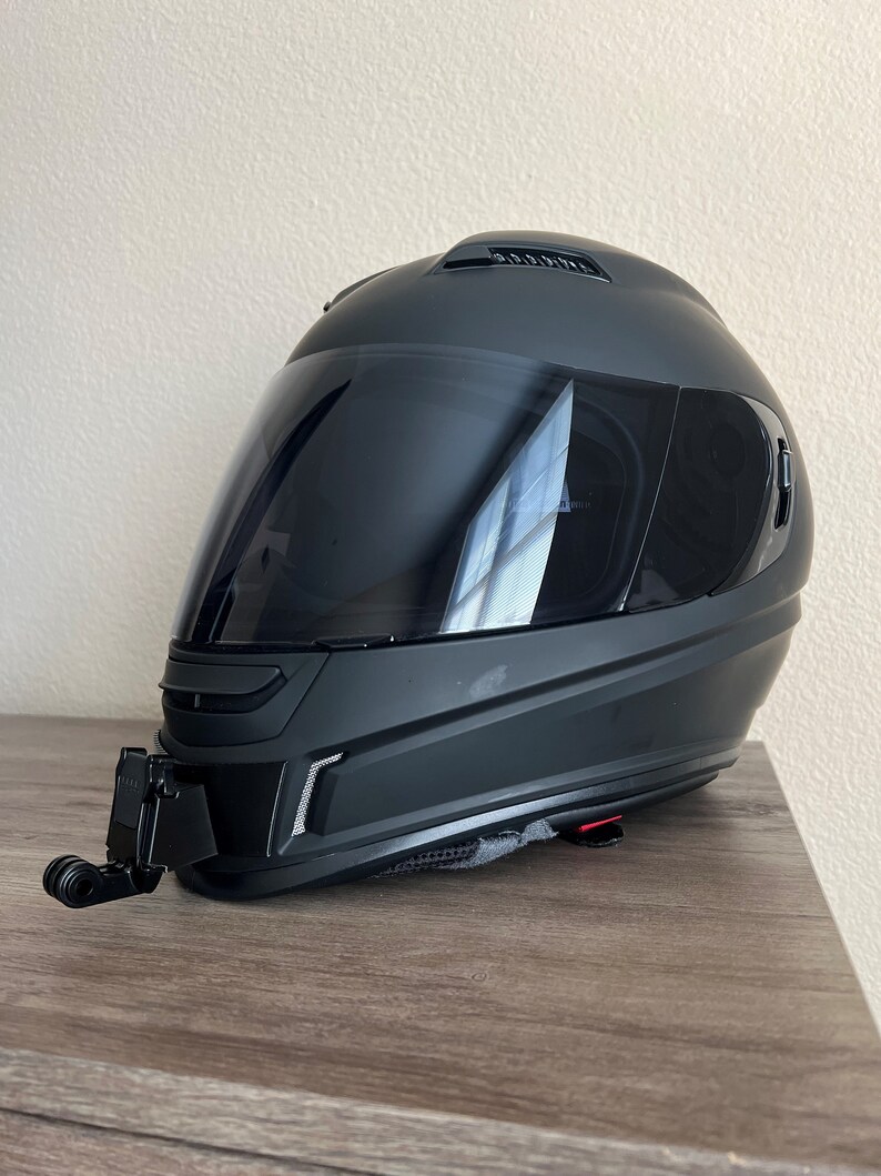 May include: A black motorcycle helmet with a clear visor and a black camera mount attached to the bottom. The helmet is sitting on a wooden surface.