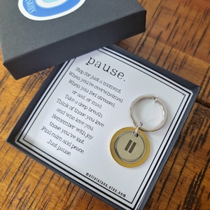 May include: A gold-colored keychain with a double vertical line symbol on a white background. The keychain is in a black box with a white card that reads "pause. Stop for just a moment. When you're overwhelmed. When you feel stressed, or sad, or mad. Take a deep breath. Think of those you love and who love you. Remember with joy those you've lost. Find calm and peace. Just pause." and the website address "mativisions.etsy.com".