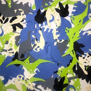 May include: A collection of dinosaur-shaped paper cutouts in various colors, including green, blue, gray, black, and white. The cutouts are in different shapes and sizes, and some are skeletons.