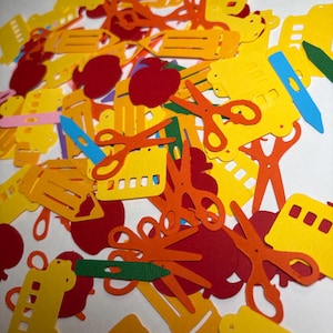 May include: A collection of school-themed paper confetti shapes in various colors including red, yellow, orange, blue, green, and pink. The shapes consist of apples, scissors, pencils, and school buses scattered across a flat surface.