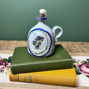 May include: A small, ceramic oil lamp with a handle, painted in shades of blue, brown, and white. The lamp features a floral design and a wick. It sits atop two vintage books with green and yellow covers.