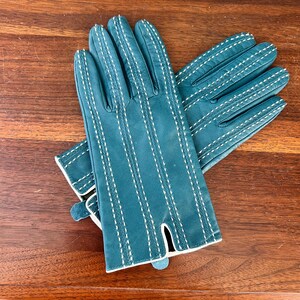 May include: Teal leather gloves with white stitching. The gloves have a classic design with vertical stitching details and a contrasting white trim around the edges. The gloves are shown on a wooden surface.