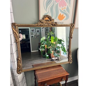 May include: A rectangular wall mirror featuring an ornate gold-toned frame with intricate scrollwork and floral carvings along the sides. The top center of the frame is adorned with a decorative crest featuring a bow and ribbon motif.