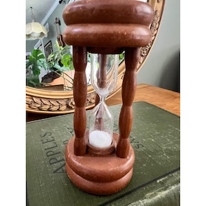May include: A wooden hourglass featuring a brown frame with a circular top and base connected by four turned pillars. The central glass bulb contains white sand, with a portion of the sand currently resting in the bottom chamber of the device.