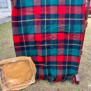 May include: A plaid blanket in red, green, navy, and yellow hangs on a wooden rack. The blanket has a fringed edge. A tan zippered bag with a clear window sits below the blanket.