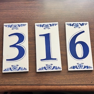 Pode incluir: Três azulejos cerâmicos retangulares com os números 3, 1 e 6 em azul. Cada azulejo tem um fundo branco com desenhos florais e de rolagem azuis na parte superior e inferior. Os números estão em negrito, com uma fonte serif.