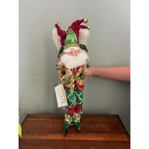 May include: A decorative elf figurine featuring a red and green jester-style hat with gold bells, a white faux fur beard, and a patterned jacket in shades of gold, red, and green. The figure wears green leggings with gold trim and has a white tag attached.