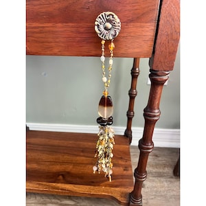 May include: A decorative pull featuring a round, ornate, antique-style metal knob attached to a long, dangling tassel. The tassel is composed of a gold-toned chain, amber-colored glass beads, and a cluster of small, multi-colored glass beads.