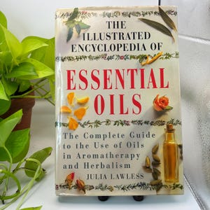 May include: A book titled "The Illustrated Encyclopedia of Essential Oils" with red lettering. The cover features botanical illustrations, including a rose, orange petals, and a small bottle of oil. The book is a guide to aromatherapy and herbalism.