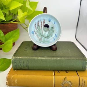 May include: A small, round, light blue ceramic dish with a floral design featuring green leaves, purple stems, and blue flowers. The dish is displayed on a wooden stand, resting on top of a stack of vintage books with green and yellow covers.