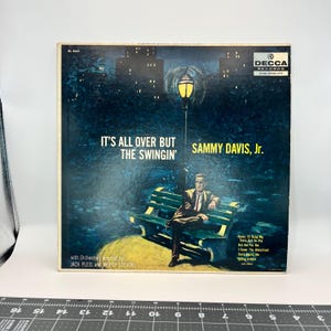 May include: Vintage vinyl record album cover for "It's All Over But The Swingin'" by Sammy Davis, Jr. The cover art features a nighttime scene with a man sitting on a park bench under a lamppost. The album is from Decca Records.