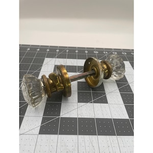 May include: A vintage door knob set featuring two clear, faceted glass knobs connected by a central metal spindle. The hardware includes aged brass circular plates and a small thumb turn mechanism, positioned on a grid mat with inch markings.