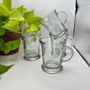 May include: Four clear glass mugs with handles, stacked on a white surface. Each mug has a light blue tree design. A green plant in a brown pot is in the background.