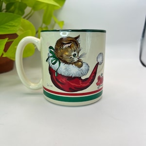 Puede incluir: Taza de cerámica color crema con asa y borde verdes, con un gatito en un gorro de Papá Noel rojo y blanco. La taza tiene una franja verde y roja cerca de la base y una pequeña ilustración de ratón.