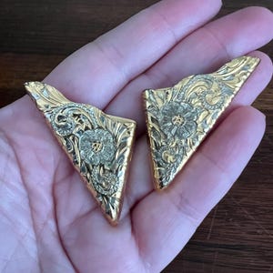 Può includere: Due orecchini dorati a forma triangolare con disegni floreali e a volute. Gli orecchini hanno una finitura lucida e sono probabilmente in metallo. I dettagli intricati dei motivi floreali sono visibili.