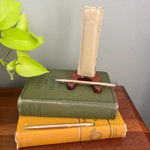 Könnte beinhalten: Ein goldfarbener Stift und das passende Etui werden auf einem Holzständer präsentiert, der auf zwei alten Büchern ruht. Die Bücher sind grün und gelb, mit den Titeln "Apples and Honey" sichtbar. Der Stift ist schlank und elegant.
