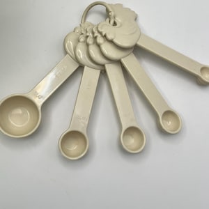 May include: A set of off-white measuring spoons, connected by a ring with a rooster-shaped design. Each spoon is labeled with a measurement, including 1/4 tsp, 1/2 tsp, 1 tsp, 1 tbsp, and 1/2 tbsp. The spoons are made of a smooth, durable plastic.