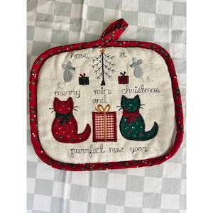 Puede incluir: Un agarrador de tela cuadrado con borde rojo que presenta bordados de gatos, ratones, regalos y un árbol de Navidad. El texto dice: have a merry mice and christmas and purrfect new year. Incluye un lazo para colgar en la parte superior.