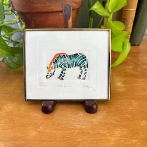 May include: A small framed print featuring a watercolor illustration of a zebra with blue and black stripes and an orange mane. The artwork is signed and titled Cebra Padilla 92 with the edition number 12/100 written in pencil below the image.