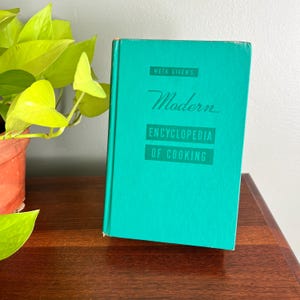 May include: A teal-colored cookbook titled "Modern Encyclopedia of Cooking" by Meta Given. The book is displayed on a wooden surface with a potted plant in the background. The book's title is in a white, stylized font.