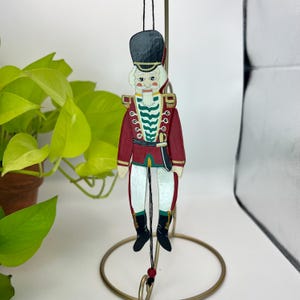 May include: A wooden Christmas ornament in the shape of a nutcracker soldier. The ornament is painted with a red jacket, white pants, and black boots. The soldier has a white beard and a black hat. The ornament hangs from a black string.