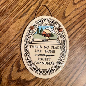 May include: Oval decorative plaque with the text "THERE'S NO PLACE LIKE HOME EXCEPT GRANDMA'S". The plaque features a painted scene of a house and landscape viewed through a window, with a brown string for hanging.