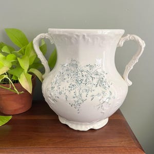May include: A white ceramic pitcher with two ornate handles and a floral design in teal. The pitcher has a wide base and a flared top, and is likely used for decorative purposes.