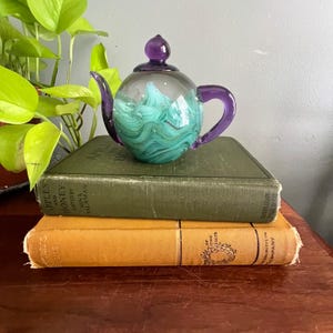 May include: A glass teapot featuring a clear body with swirling teal and white patterns inside, complemented by a solid purple handle, spout, and lid. The teapot is resting on top of two stacked hardcover books, one green and one yellow.