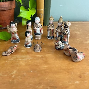 May include: A collection of small, hand-painted clay figurines depicting a nativity scene. The set includes figures of people in robes, an angel with gold wings, a central infant in a manger, and several small animals including sheep and a donkey.