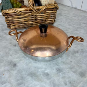 Puede incluir: Una olla de cocina de cobre y plata con un pomo negro en la tapa. La olla tiene asas en forma de lazo y una forma redondeada. Una cesta de mimbre llena de comida está en el fondo.