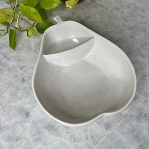 May include: A speckled white pear-shaped serving dish with a small, raised compartment. The dish has a stem-like handle and is designed for serving snacks or appetizers. The overall design is simple and elegant.