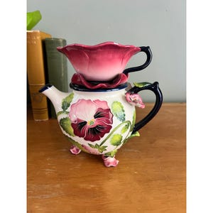 Może przedstawiać: Ceramiczny zestaw do herbaty dla jednej osoby, składający się z białego czajniczka z ręcznie malowanym wzorem bratków w odcieniach różu i fioletu, zielonymi liśćmi oraz ciemnoniebieskim uchwytem. Pasująca różowa filiżanka w kształcie kwiatu stoi na górze.
