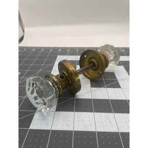 May include: A vintage door knob set featuring two clear, faceted glass knobs attached to aged brass circular plates and a central metal spindle. The hardware displays a weathered patina on the metal components, contrasting with the transparent glass.
