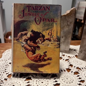 May include: Vintage book titled "Tarzan and the Jewels of Opar" by Edgar Rice Burroughs. The cover features a dynamic illustration of Tarzan battling a lion-like creature. The book has a worn, aged appearance, suggesting it is an antique.