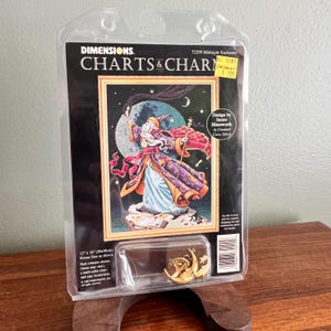 May include: A Dimensions Charts & Charms cross-stitch kit featuring a wizard with a staff, moon, and stars. The kit is in a clear plastic package with a gold-colored pin. The frame size is 12 x 18 inches.