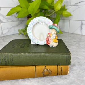 Puede incluir: Un marco de fotos de cerámica blanca con una abertura redonda, decorado con diseños florales y de vid pastel. Una pequeña figura de un conejo con sombrero y abrigo está adjunta. El marco se coloca encima de dos libros antiguos.