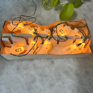 May include: A string of Halloween ghost lights in a cardboard box. Each ghost is white with a smiling face and a small light bulb inside, emitting a warm orange glow. The lights are connected by a black wire.