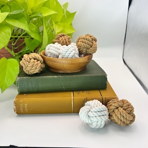 May include: A collection of decorative rope knots in shades of tan and white. Some knots are in a wooden bowl, others are placed on top of vintage books with green and yellow covers. A houseplant is in the background.