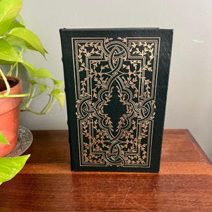 May include: A dark green hardcover book featuring an intricate gold foil stamped design on the front cover. The pattern consists of geometric interlacing lines and delicate leaf motifs arranged in a symmetrical, rectangular frame.
