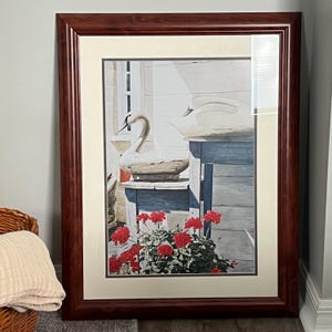 May include: Framed artwork featuring a watercolor painting of two white swans. The swans are positioned near a blue table and a window. Red flowers are in the foreground. The frame is a dark brown color.
