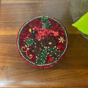May include: A round, burgundy velvet box with an embroidered design. The design features a red bird with sequin details, a brown pinecone, and green beaded foliage. The box is trimmed with gold and green beads.