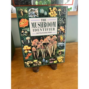 May include: A hardcover book titled The Mushroom Identifier by David Pegler and Brian Spooner. The cover features a collage of various mushroom species in natural settings, with a central image of a cluster of brown-capped mushrooms.