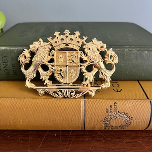 Puede incluir: Broche dorado con un escudo heráldico con una corona y dos leones. El escudo tiene un diseño de escudo con cuatro cuadrantes, cada uno con un emblema diferente. El broche está sobre libros antiguos.