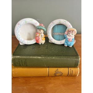 May include: Two circular ceramic picture frames featuring decorative floral vines. One frame features a rabbit figure wearing a green hat and yellow coat. The other frame features a kitten figure in a blue outfit with the text BEATRIX POTTER visible.