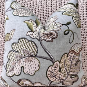 Peut inclure: Un coussin bleu clair avec un motif floral dans les tons de vert, marron et blanc. Le coussin pr&eacute;sente un motif r&eacute;p&eacute;titif de feuilles et de fleurs stylis&eacute;es.