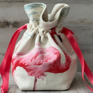 May include: A drawstring bag featuring a pink flamingo design on a cream-colored fabric. The bag has a pink ribbon closure and a striped pattern at the top. The bag is made of a textured fabric.