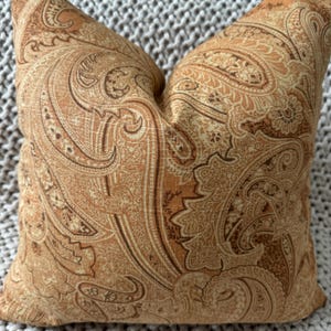 May include: A decorative square pillow with a warm color palette. The pillow features a detailed paisley pattern in shades of brown, tan, and gold. The fabric has a slightly textured appearance, adding depth to the design. The pillow is likely used for home decor.