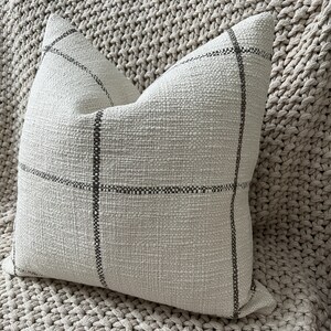 May include: A cream-colored pillow with a subtle black and white checkered pattern. The pillow is made of a textured fabric and has a simple, modern design.