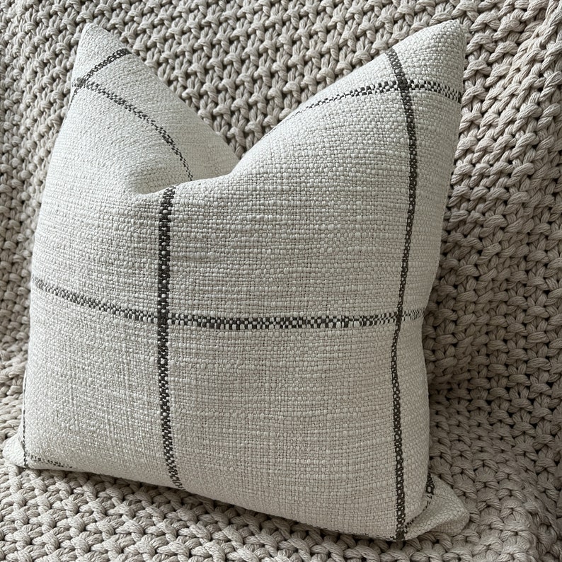 May include: A cream-colored pillow with a subtle black and white checkered pattern. The pillow is made of a textured fabric and has a simple, modern design.